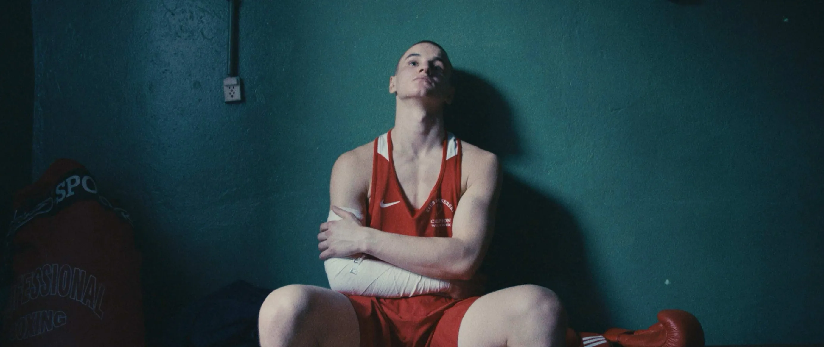 Łukasz in a red boxing vest sits against a teal wall, arms folded, head tilted back
