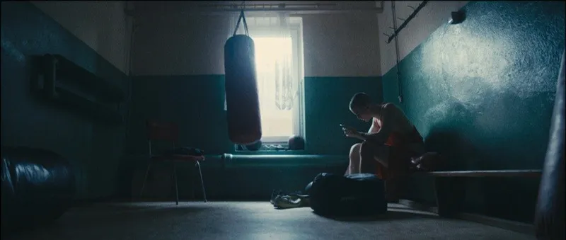 A lone boxer sits beneath a hanging punching bag in an empty gym