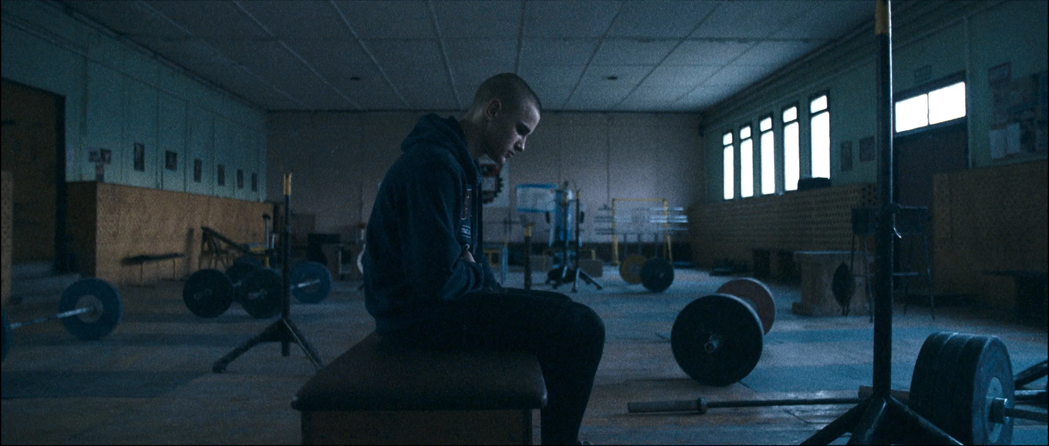 Łukasz sits alone on a bench in an empty weightlifting gym, head bowed