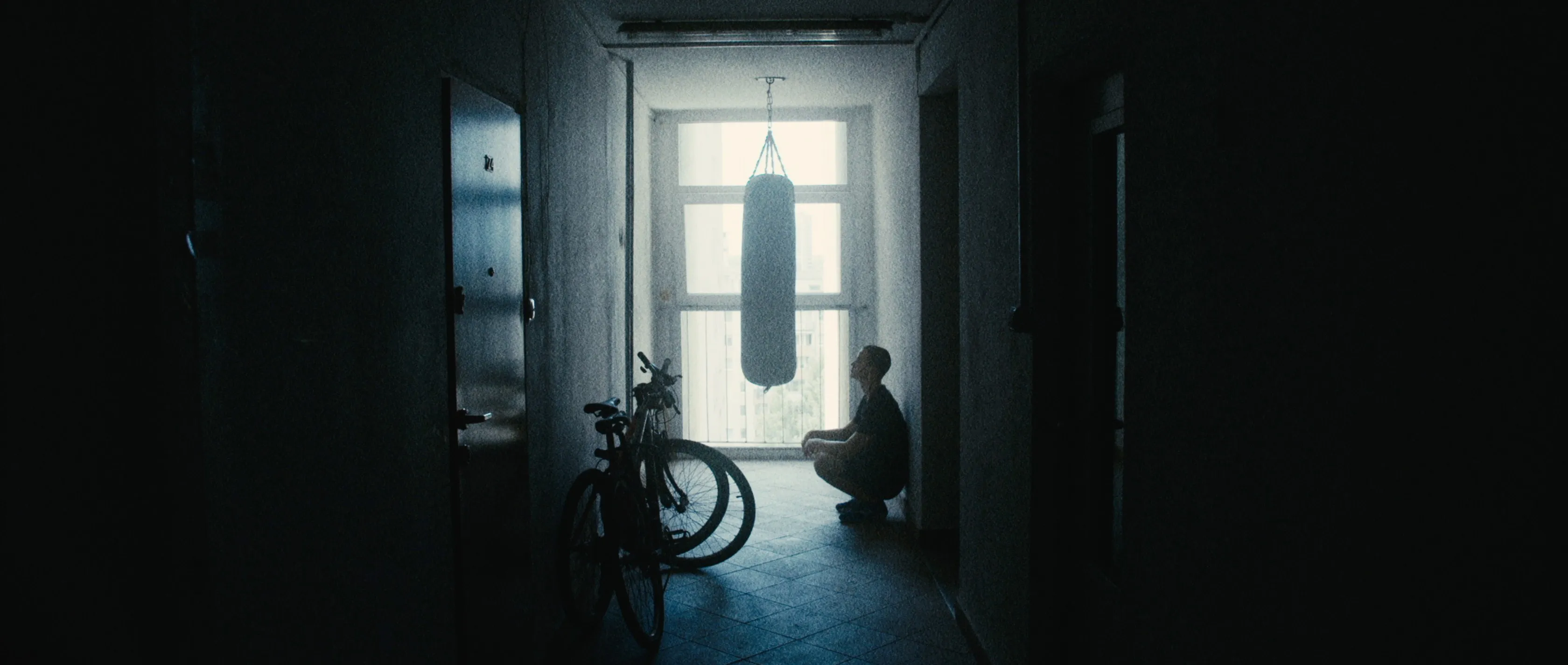 A boy crouches in a dark stairwell, looking up at a punching bag hanging in front of a bright window