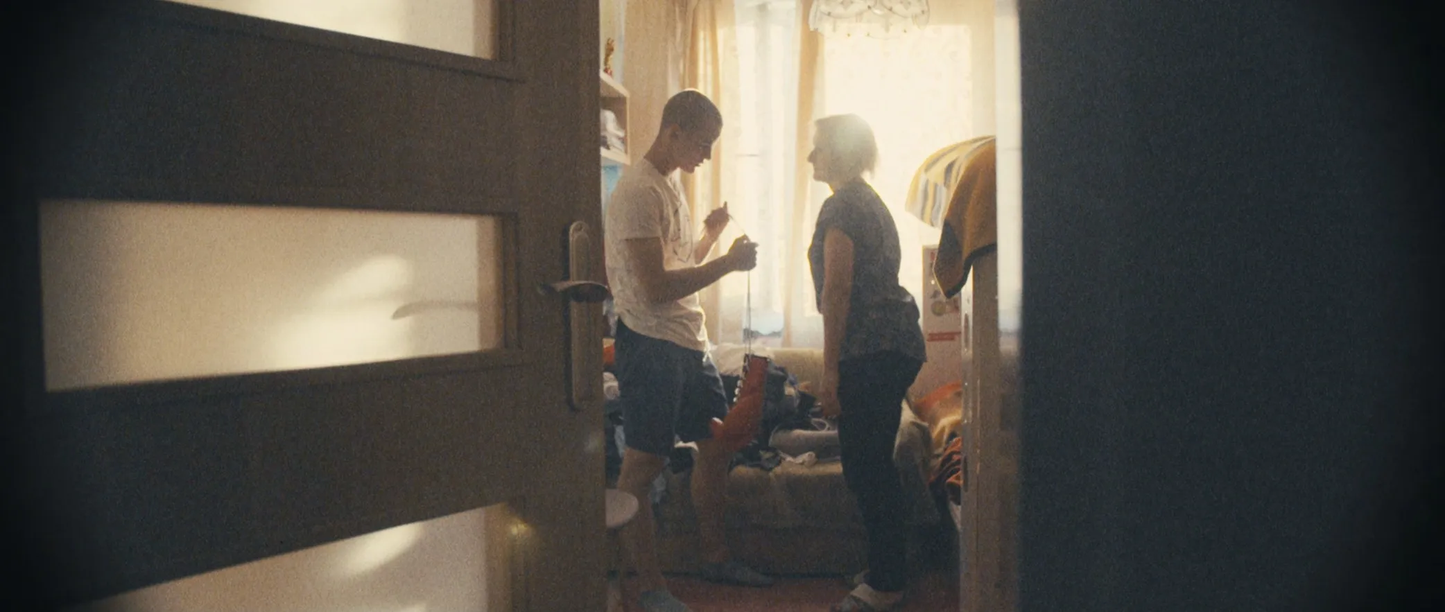Łukasz and his mother talk in a sunlit room, seen through a half-open door