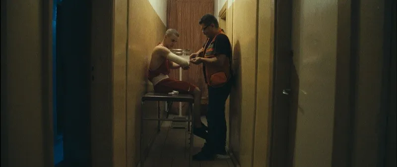A boxer sits on a table while a man prepares his hand wraps in a narrow room